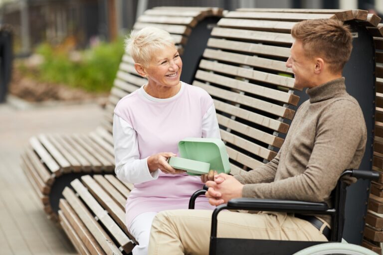 Jolly attractive mature lady with short hair sitting on bench outdoors and opening lunch box for handicapped man in wheelchair