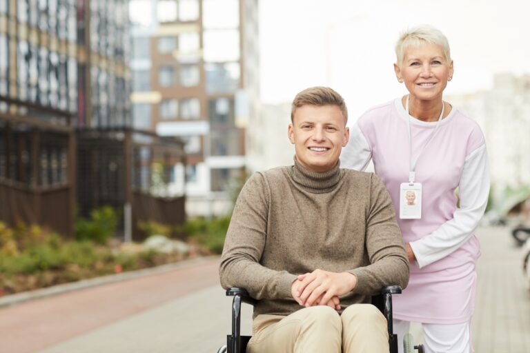 Portrait of smiling careful mature female recovery specialist with badge on neck strolling with handicapped man in wheelchair