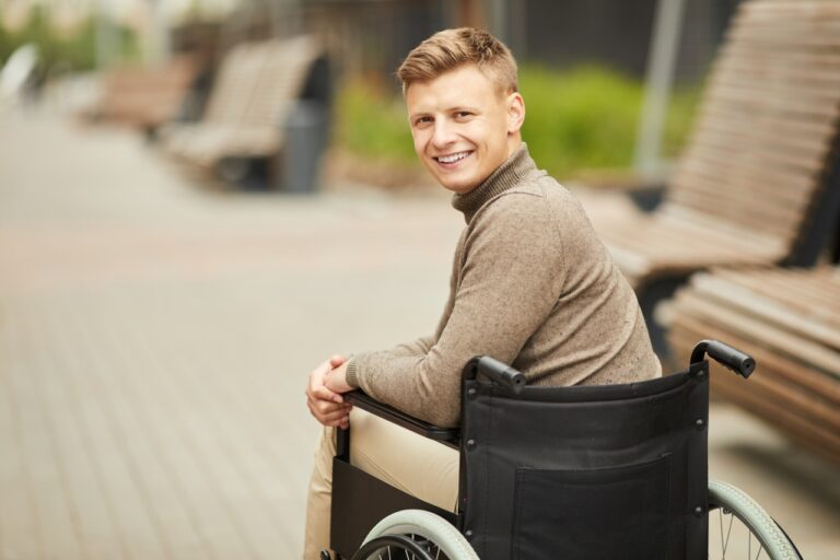 Portrait of smiling handsome young handicapped man in brown sweater sitting in wheelchair outdoors